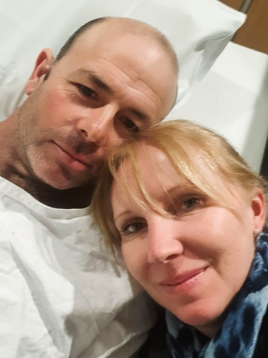 Selfie of man in hospital bed and woman leaning her head on his shoulder, a bittersweet expression on their faces.