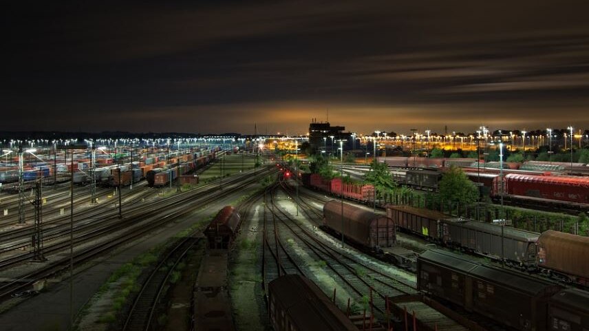 Photo of freight train station at night, there are multiple rail tracks and large red containers 