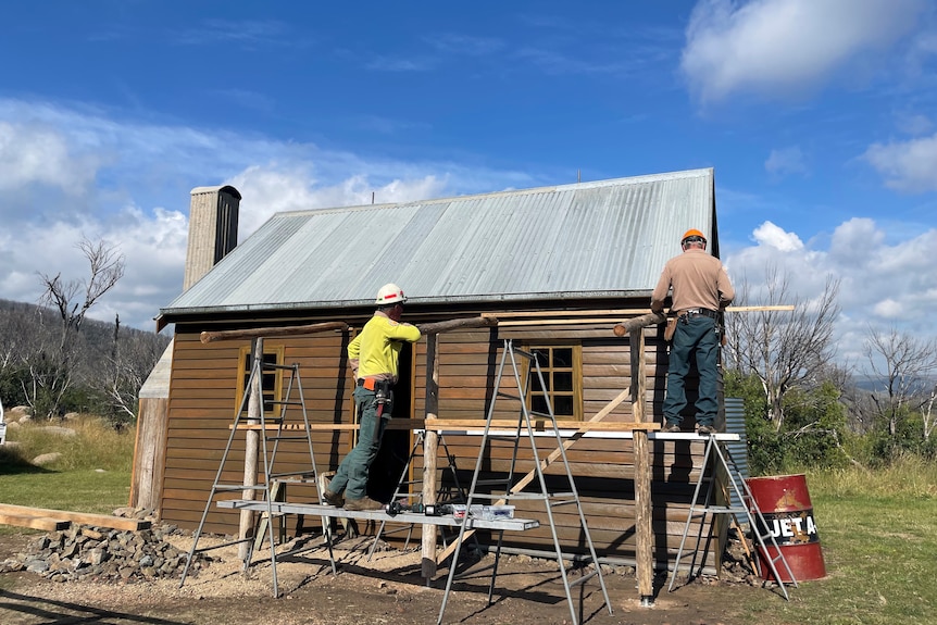 Two men in hardhats stand on scaffolding while working on a wooden hut