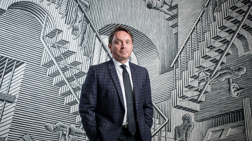 Man in navy checked suit standing in front of blown up version of M.C. Escher’s “Relativity”.