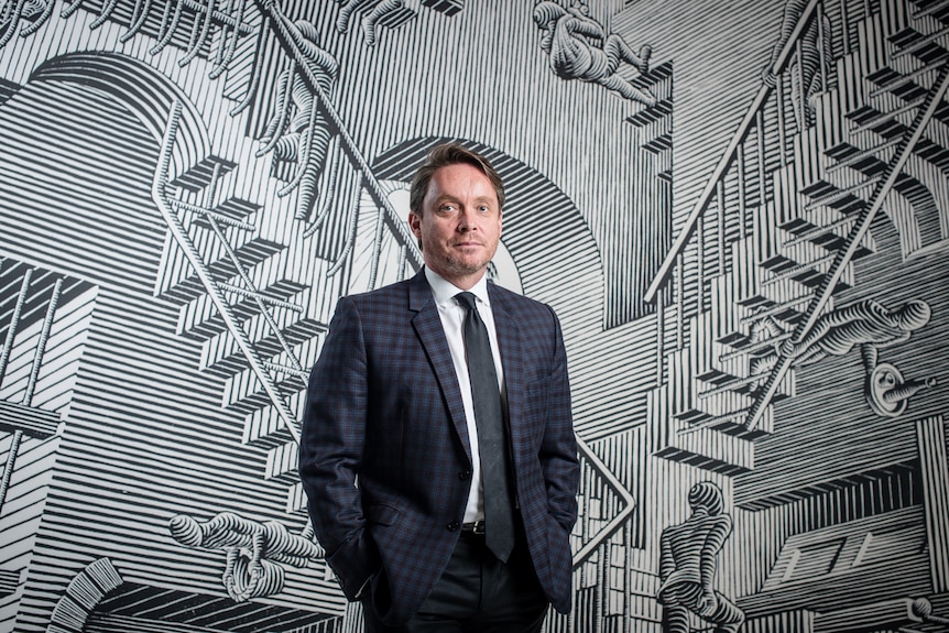 A man, wearing a suit, stands in front of a large black-and-white line drawing