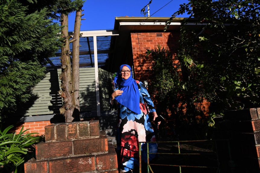 A woman wearing glasses, an electric blue headscark and bright long-sleeve dress stands outisde a single-story bbrick home