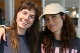 two women with long brown hair, arm in arm and smiling. one wears a light brown baseball cap 
