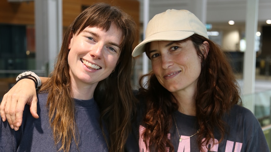 two women with long brown hair, arm in arm and smiling. one wears a light brown baseball cap 