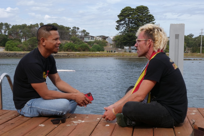 A man with dyed blonde hair sits on a wharf with legs crossed facing another man with dark curly hair, talking. 