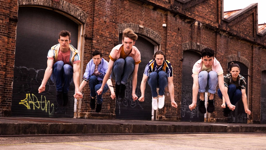 Six dancers jump in the air snapping their fingers in front of an old brick warehouse.