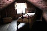 A timber clad attic room with a single iron-framed bed and a small window letting some light into the shadowy room.