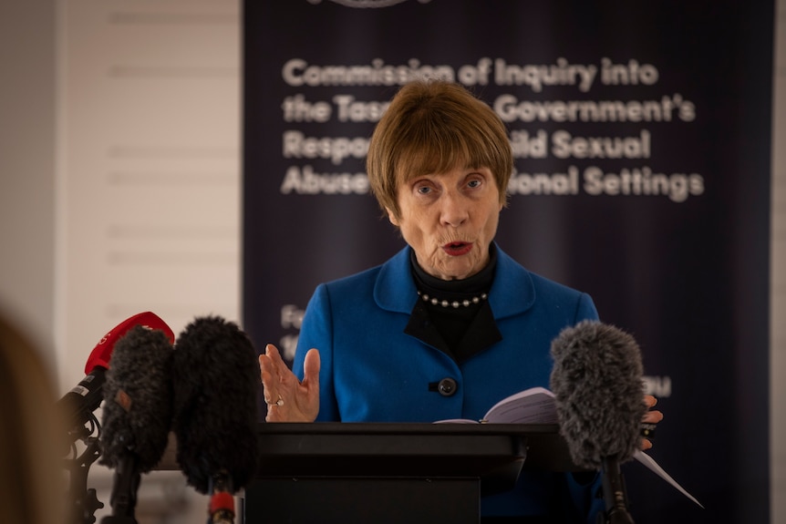 Older woman stands at microphoned podium, addressing the media 