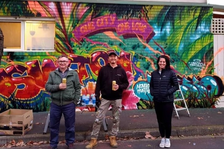 Three people with beers in their hands standing in front of a brick wall with art painted on it.
