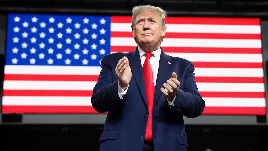 President Donald Trump is about to clap, the American flag is projected on a screen behind him