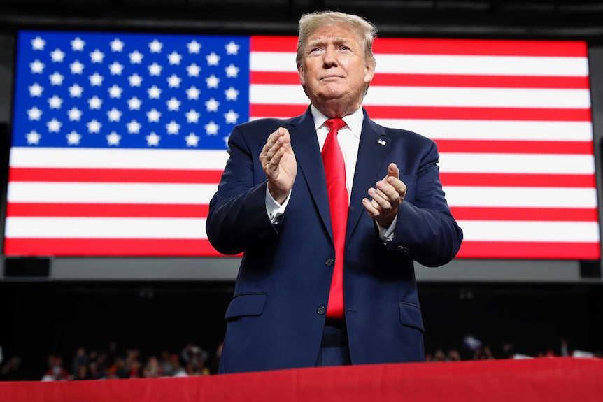 President Donald Trump is about to clap, the American flag is projected on a screen behind him