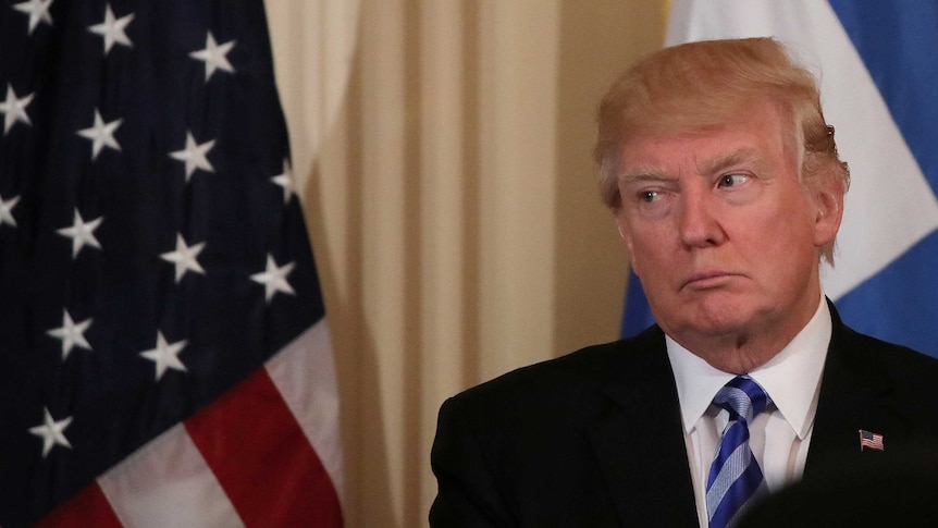 Donald Trump sternly looks to the left as he stands in front of an American and Greek flag.