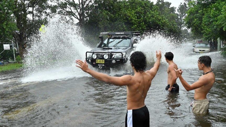 A car drives through flooded water, as a group of teenage boys watch on.