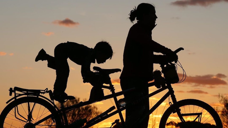 A woman stands at the front of her bike while her son plays at the end. They are silhouetted against an evening sky