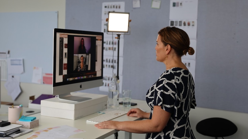 A woman with brown hair standing at a desk and facetiming on a computer, vision border behind her