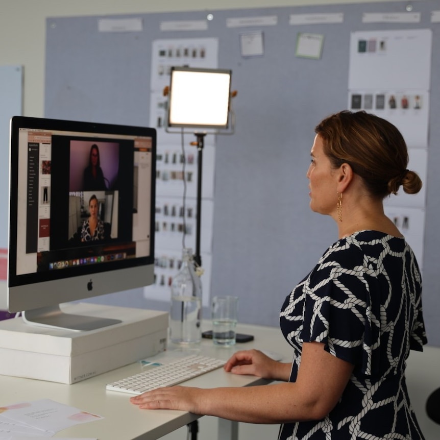 A woman with brown hair standing at a desk and facetiming on a computer, vision border behind her