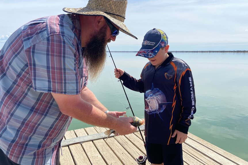 A man with a checked shirt, beard and straw hat holds a fish as a boy with a long sleeve top and cap watches on a jetty.