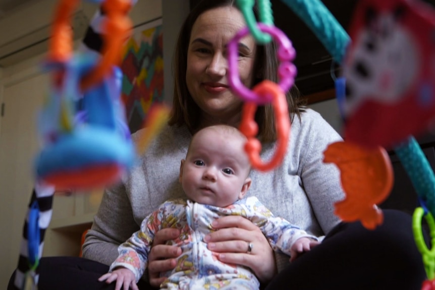 A lady with long dark hair holds a baby who is pictured through toys