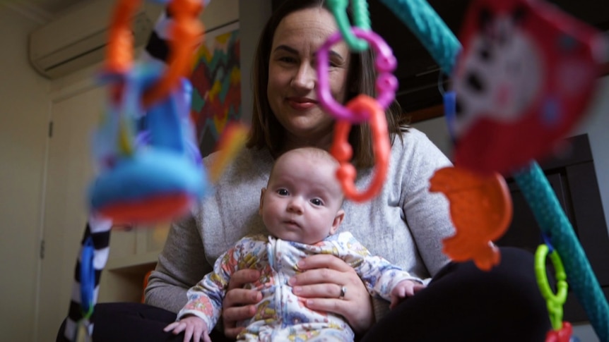 A lady with long dark hair holds a baby who is pictured through toys