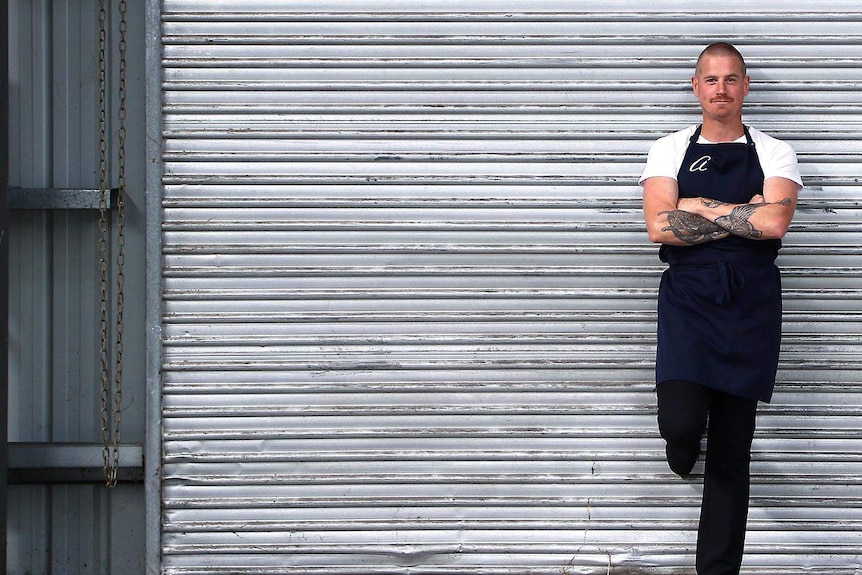 man in white t-shirt with blue apron with 'a' on it stands in front of roller door with arms crossed