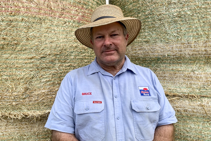 A man wearing a blue collared shirt and a hat stands in front of hay bales.