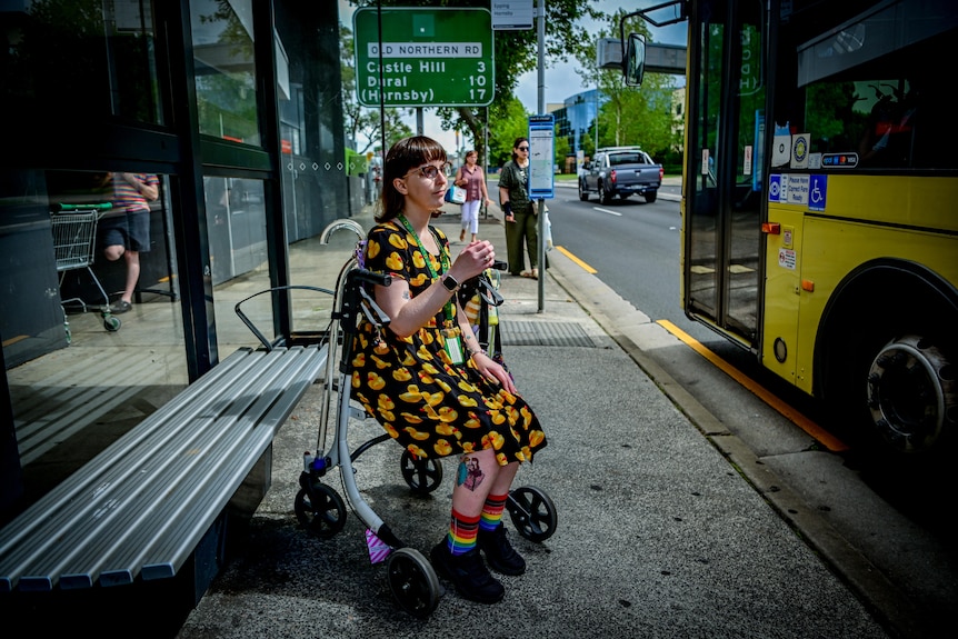 Una mujer joven con un vestido colorido sentada en una parada de autobús.  Ella tiene cabello castaño oscuro y gafas.