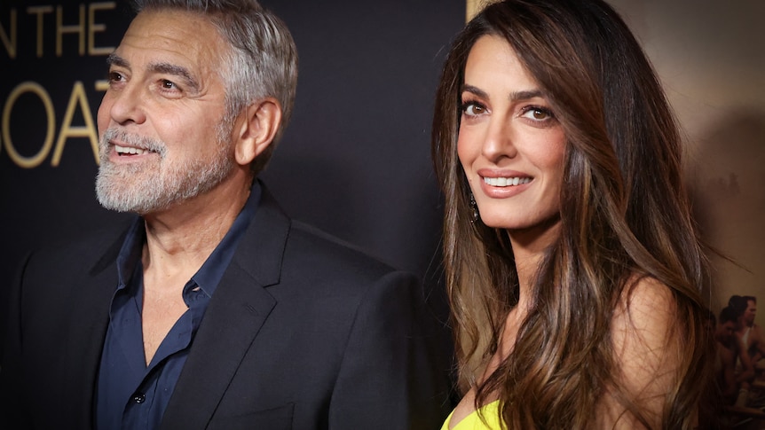 George Clooney with a grey beard smiles off camera while his wife Amal Clooney with long brown hair smiles.