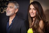 George Clooney with a grey beard smiles off camera while his wife Amal Clooney with long brown hair smiles.