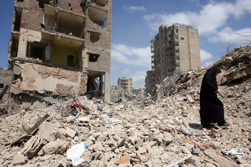 A woman wearing a long black gown walks over piles of rubble in front of two partially destroyed apartment buildings