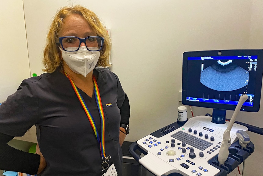 A woman wearing a mask stands next to a computer screen.