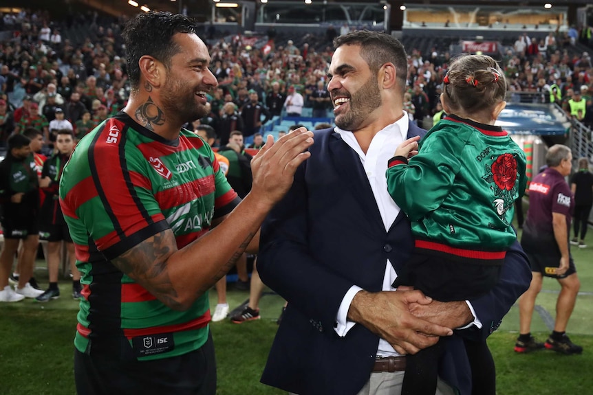 A man in a red and green Rabbitoh's uniform laughs with a man in a suit who is holding a girl. They are in crowded stadium