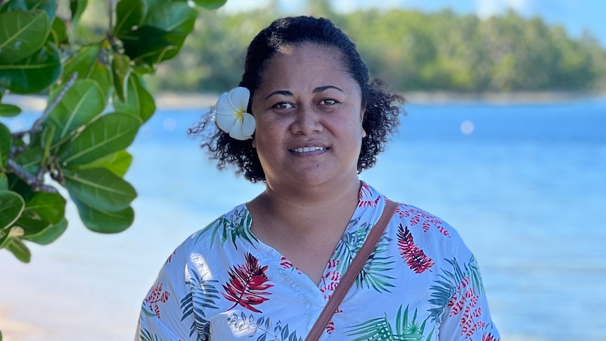 A woman wearing a floral top stands on the waters edge beside a green leafy tree on a bright sunny day. 