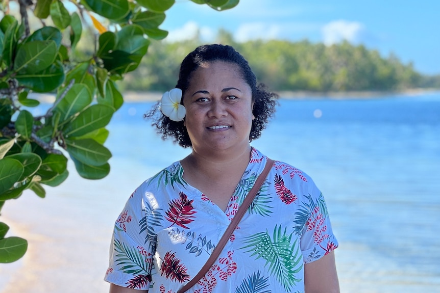 A woman wearing a floral top stands on the waters edge beside a green leafy tree on a bright sunny day. 