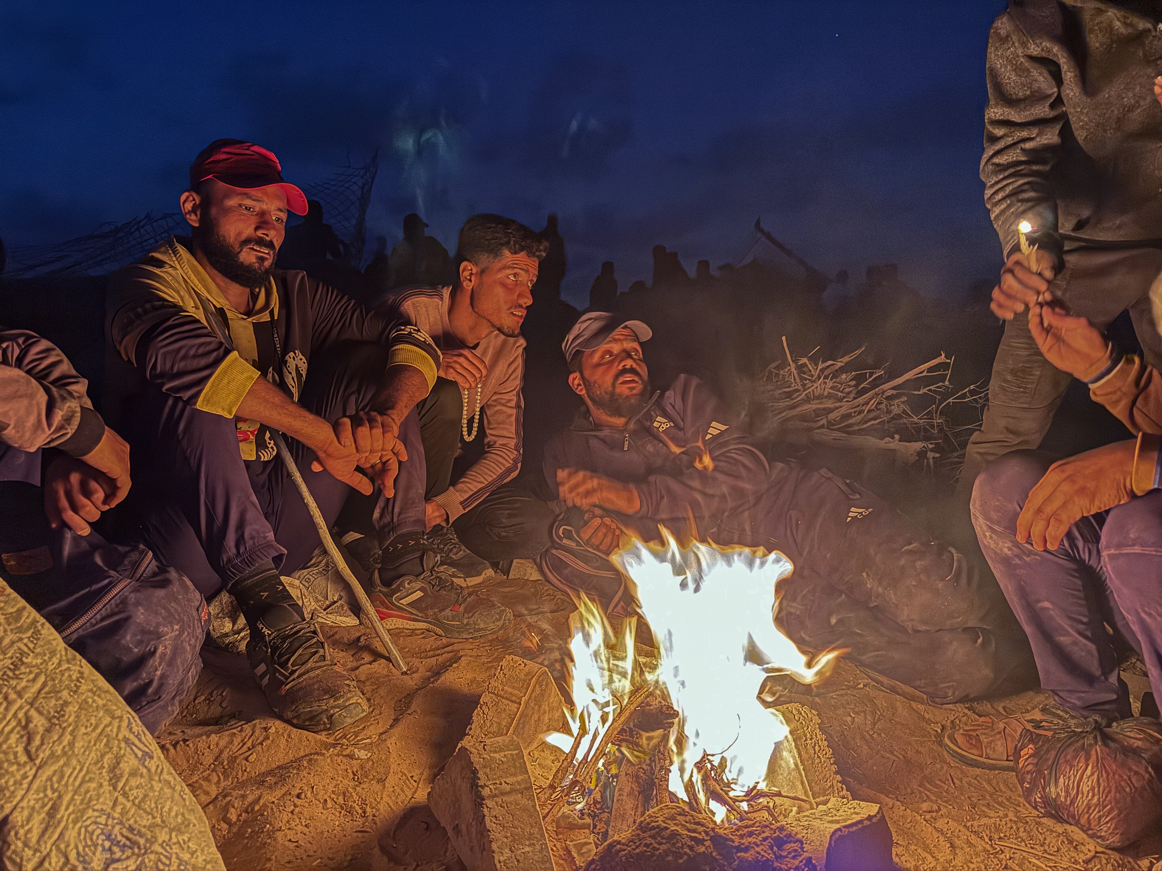 Six men sit on barren ground staring into a bright fire under a dusk sky.