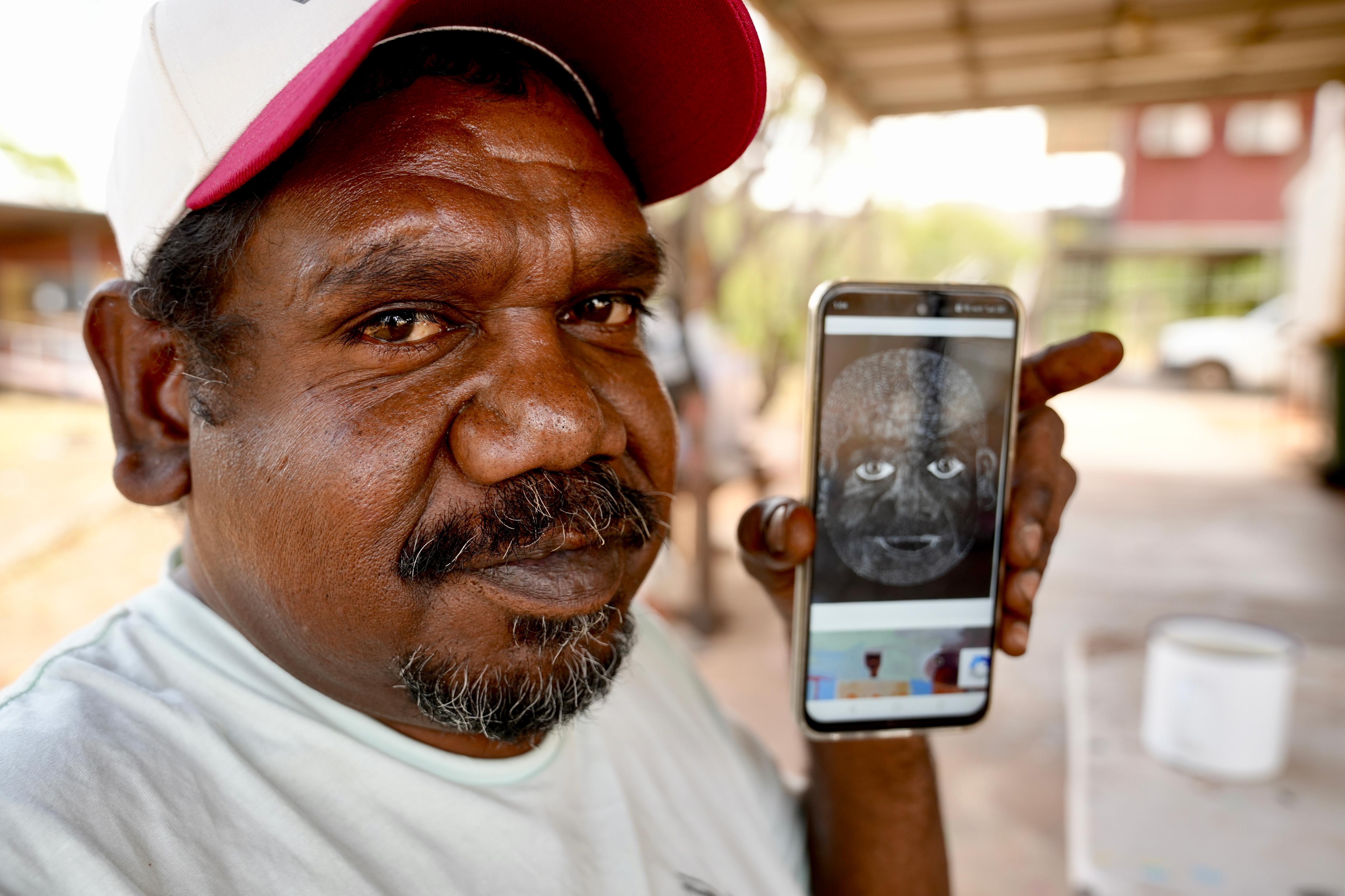 Aboriginal man wearing a cap showing a pic of his art on his phone