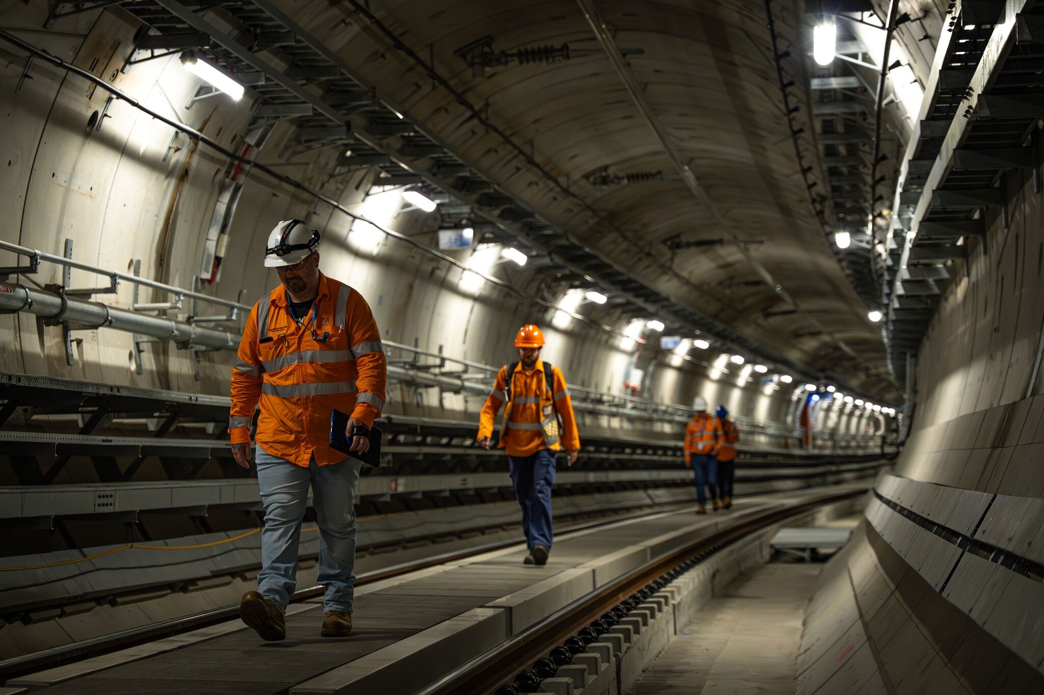 Four people in orange high vis tops, blue pants and hard hats walk along a raised concrete walkway inside a concrete tunnel.