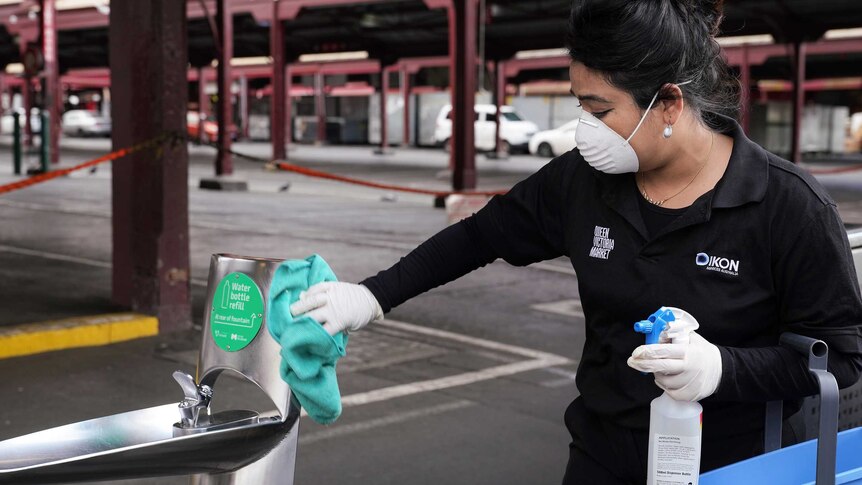 A dark-haired lady wearing dark shirt and pants, wearing white mask, wipes down water fountain at train station