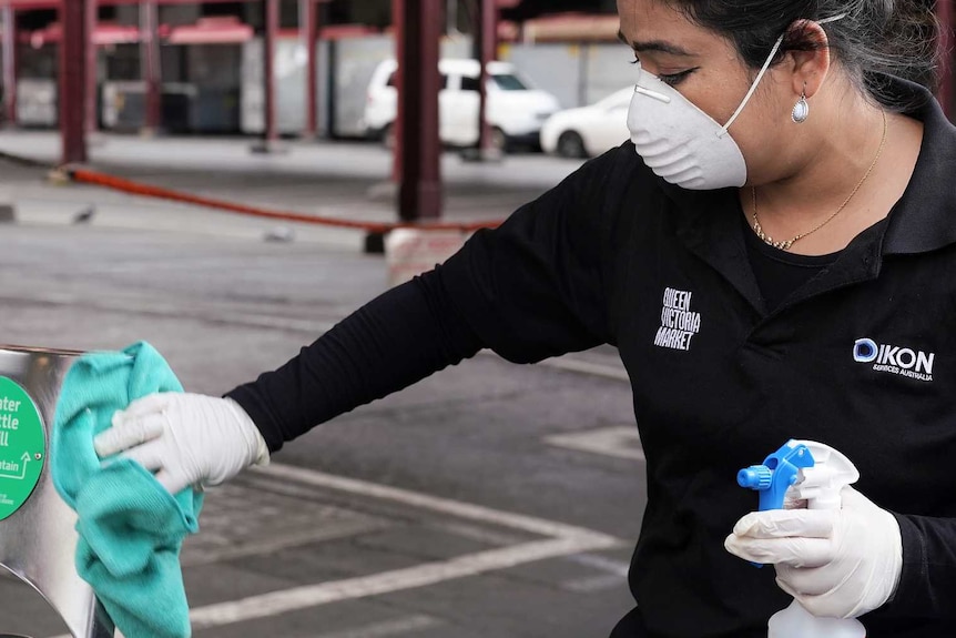 A dark-haired lady wearing dark shirt and pants, wearing white mask, wipes down water fountain at train station