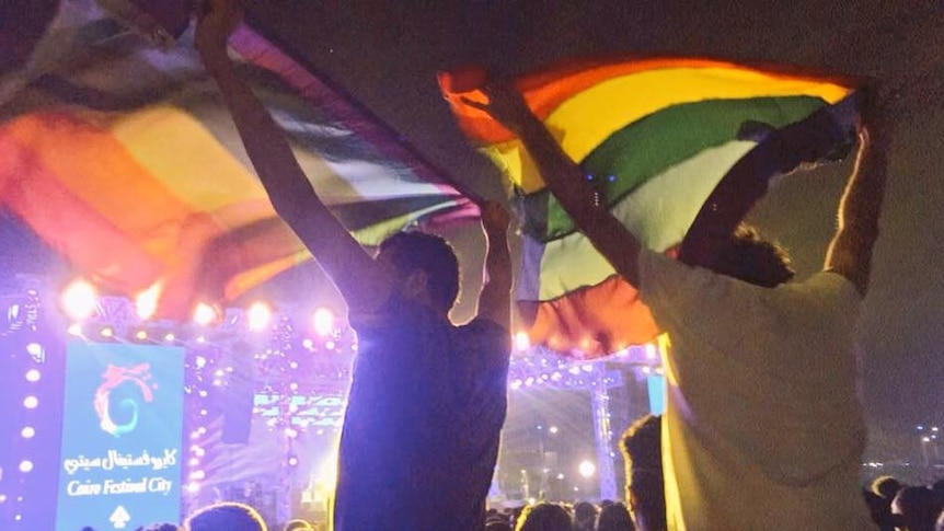 Two men sit on other peoples' shoulders and wave two rainbow flags at a concert in Cairo.