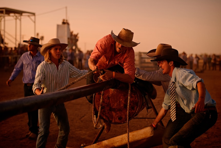 A man in a cowboy hat pulls himself along a horizontal pole using a rope, surrounded by several other people, at a rodeo course.