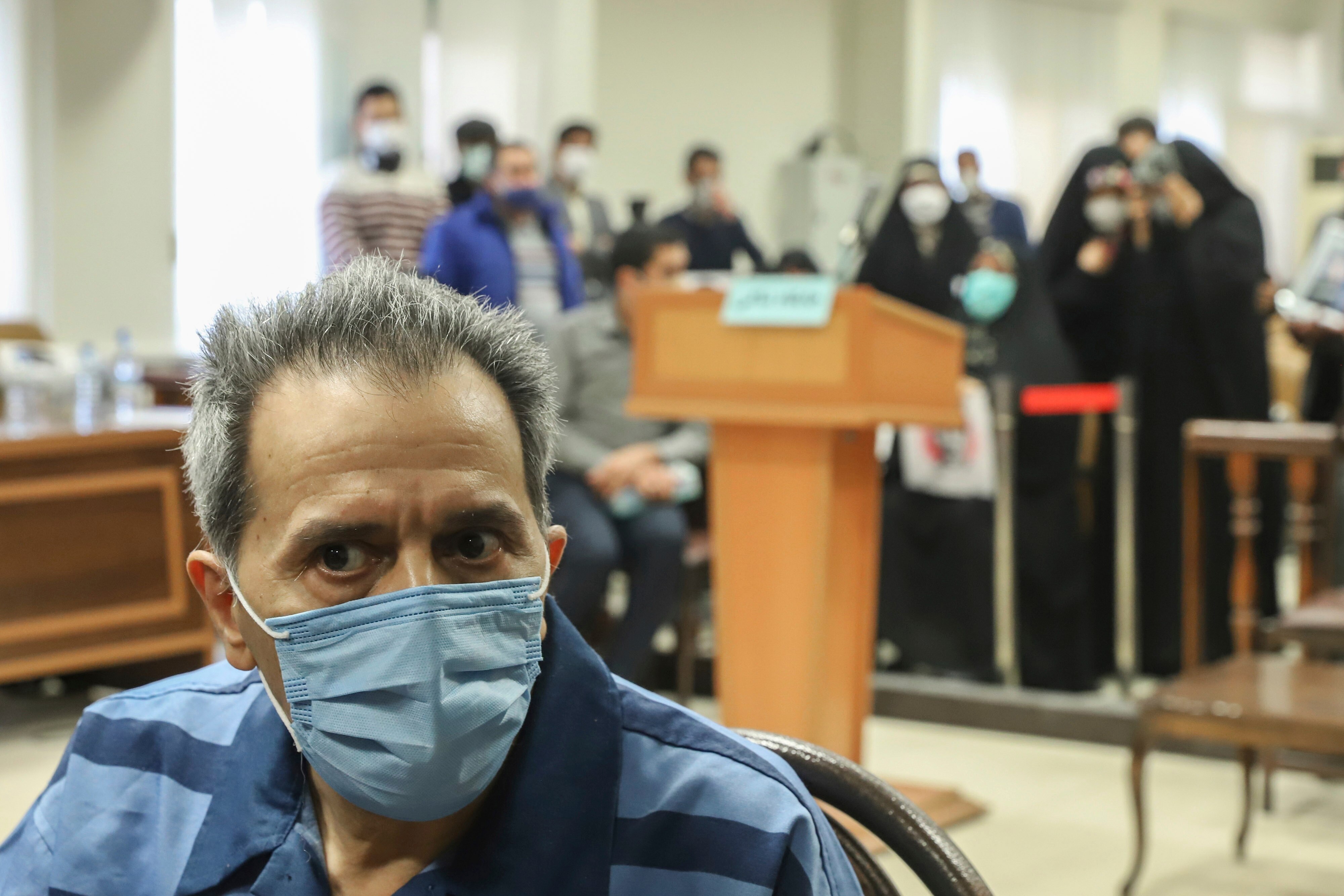 Jamshid Sharmahd wearing a blue face mask and jumpsuit while seated in a courtroom in front of a crowd of blurred supporters