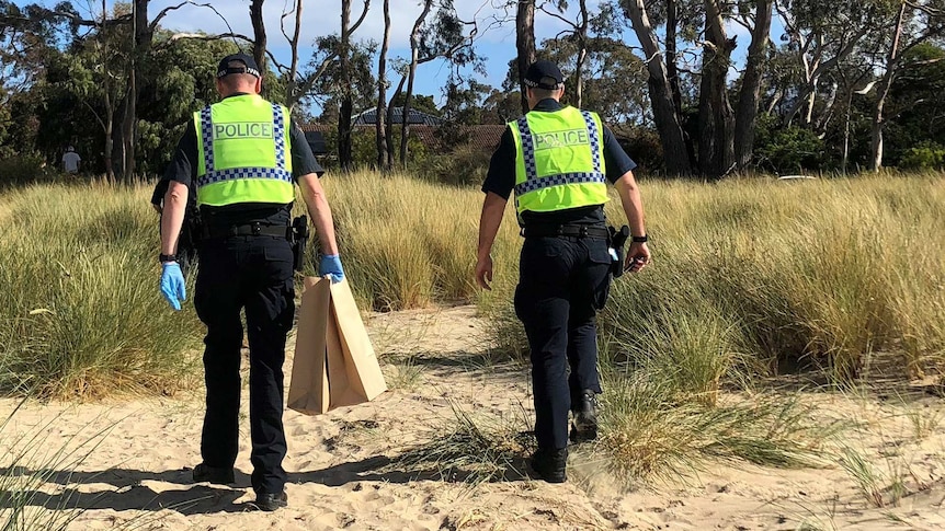 Two police officers wearing hi-vis jackets walk up off a beach through scrub. One is carrying evidence bags and wearing gloves.