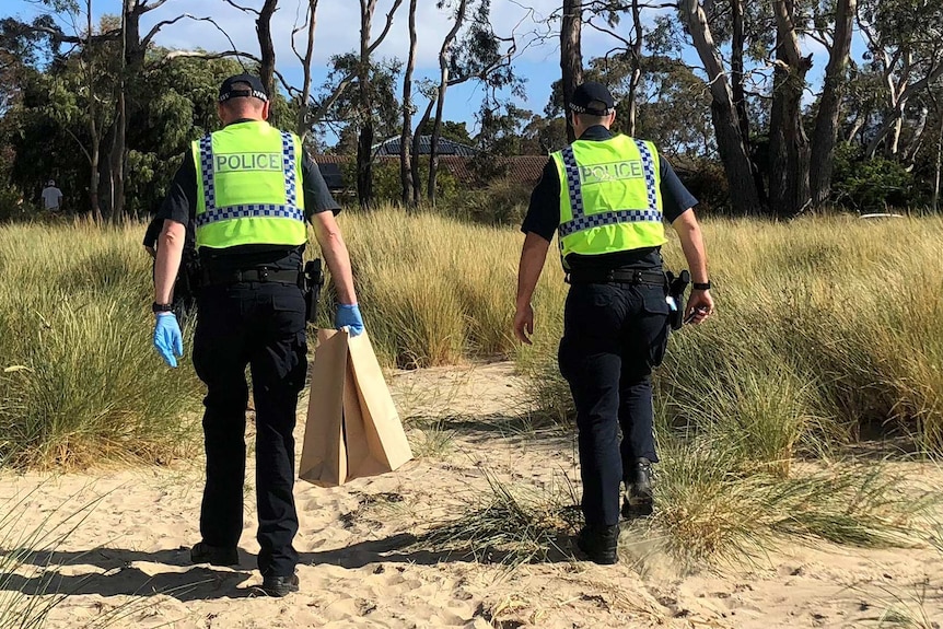 Two police officers wearing hi-vis jackets walk up off a beach through scrub. One is carrying evidence bags and wearing gloves.