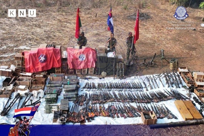 An aerial shot of three soldiers holding flags with piles of guns, ammo and equipment. 