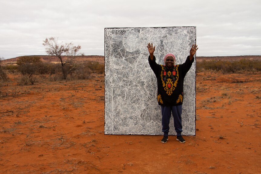 An older Aboriginal woman with grey hair, the artist Betty Muffler, stands with her hands up in front of her painting in desert