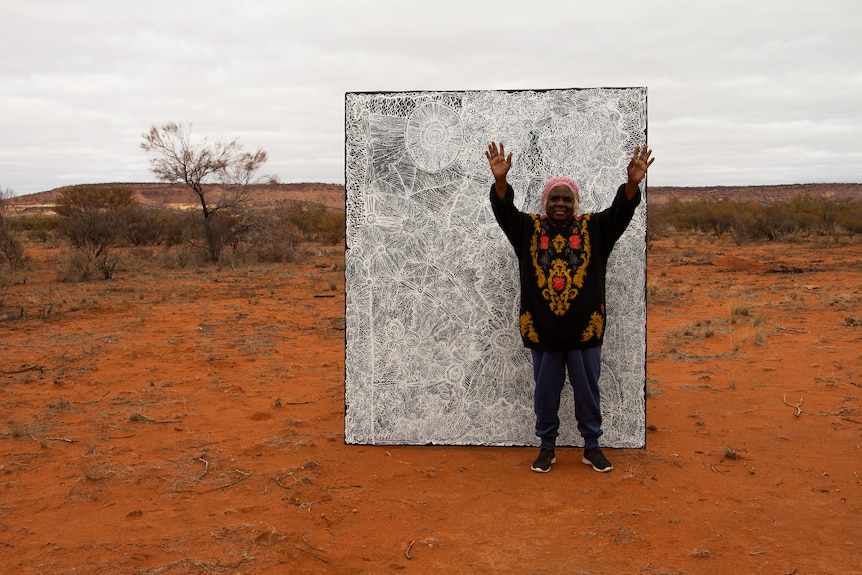 An older Aboriginal woman with grey hair, the artist Betty Muffler, stands with her hands up in front of her painting in desert