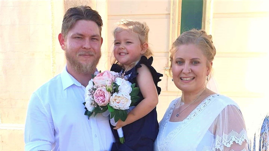 A smiling couple poses with a young girl carrying a bouquet of flowers.