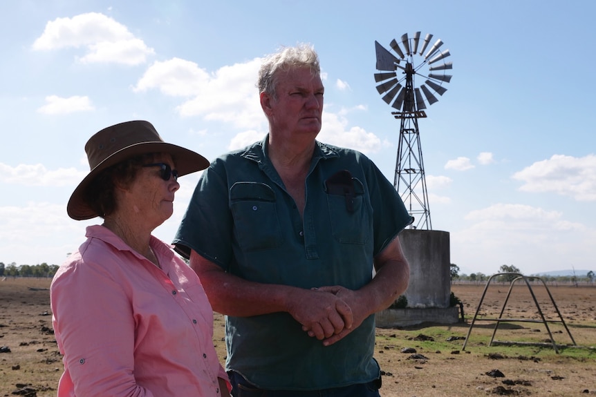 An older man and woman stand looking solemn in a paddock. A windmill is visible behind them.