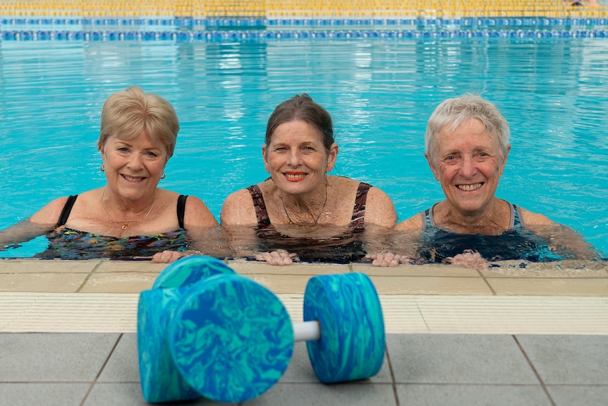 three elderly Caucasian ladies lean against the pool edge behind a set of foam dumbells.