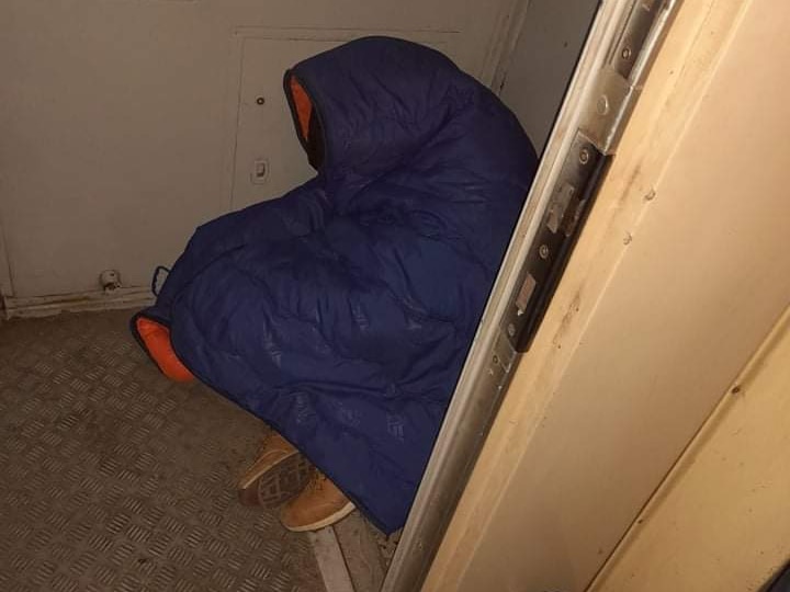 A person has a purple sleeping bag wrapped around him as he huddles in a corner.